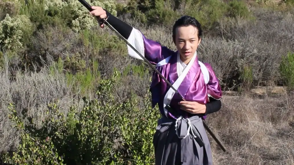 Martial artist in traditional purple and black attire holding a spear in a natural setting.